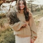 Smiling woman in cozy sweater holding a bouquet of dry flowers on a sunny outdoor path.